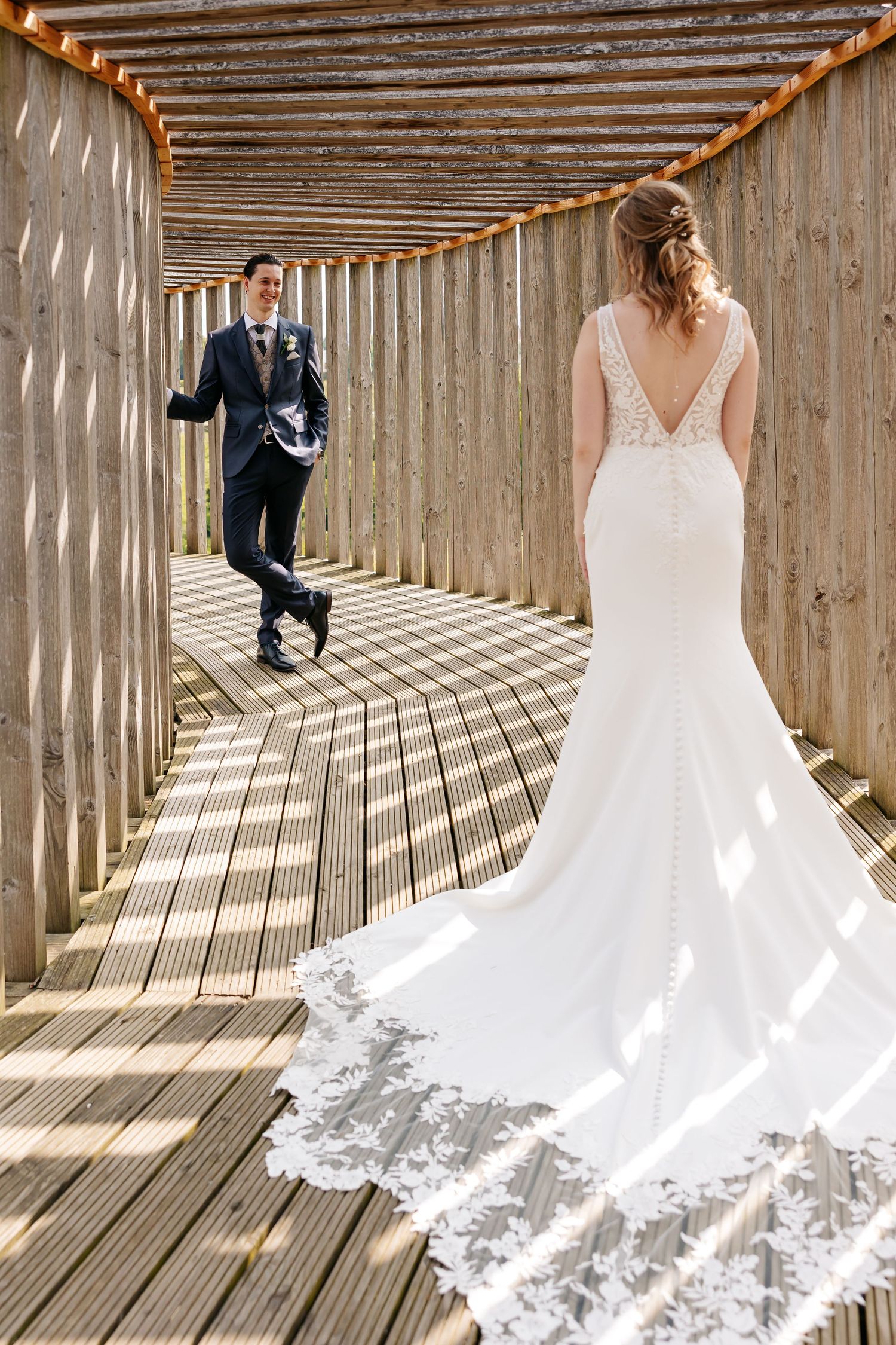Bride in lace gown with train faces groom walking toward her in wooden walkway.