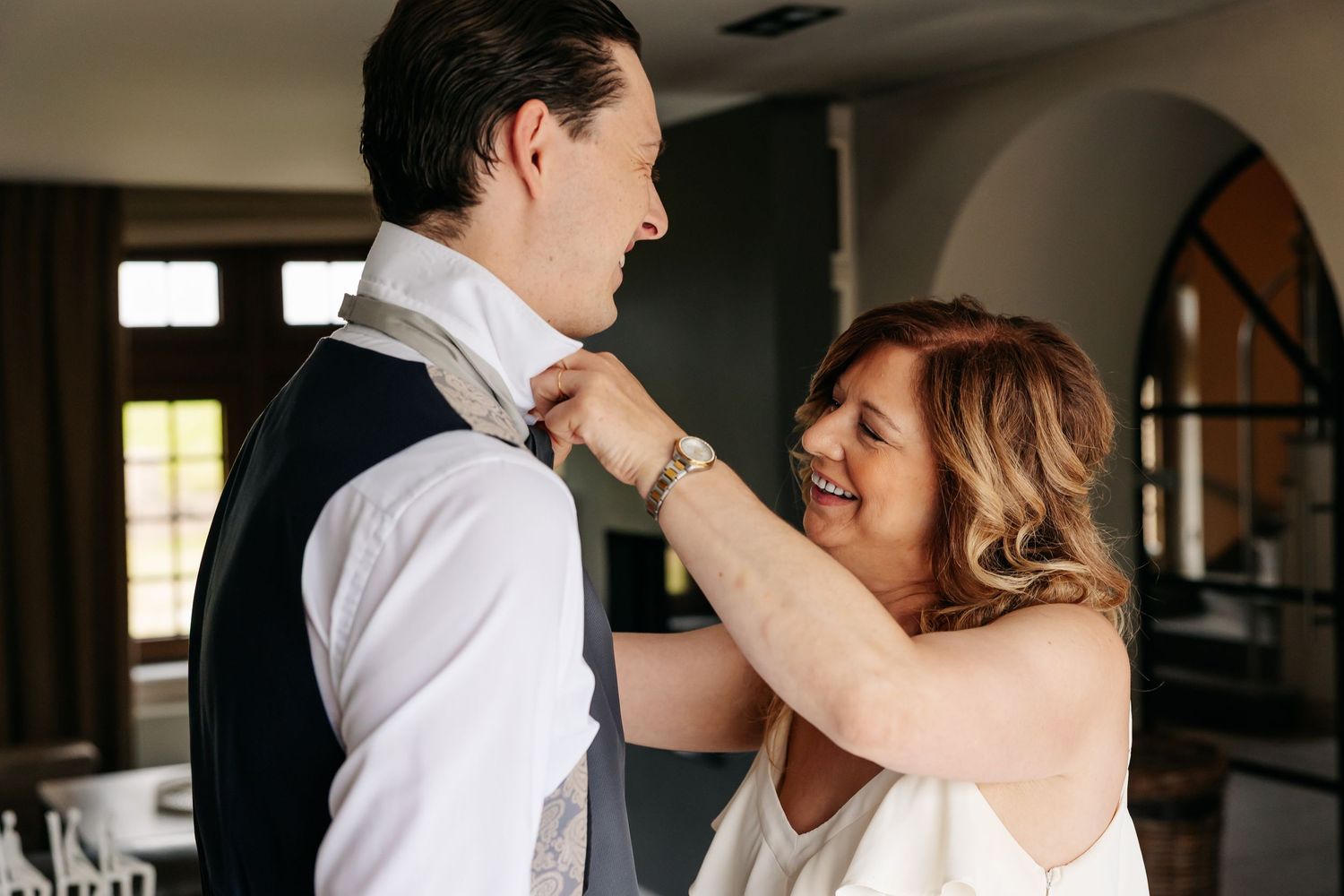 Older woman smiling while adjusting groom's tie during wedding preparation indoors.