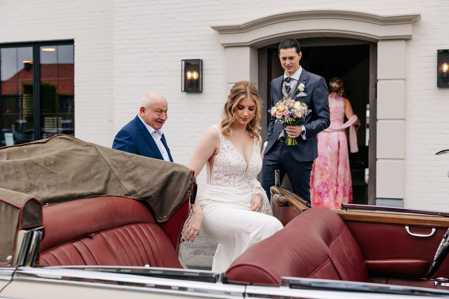 Bride in lace wedding gown steps into vintage cream convertible car assisted by older gentleman outside white venue.