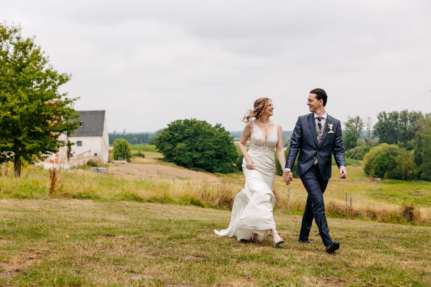 Bride in flowing white dress and groom in navy suit walking hand in hand across a green hillside.