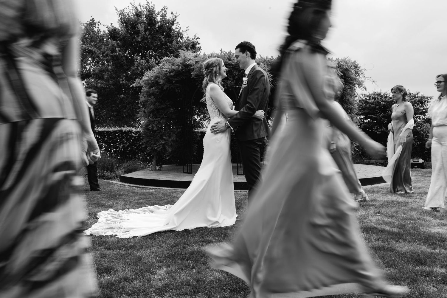 Black and white photo of bride and groom sharing a kiss while wedding guests walk past in a garden.