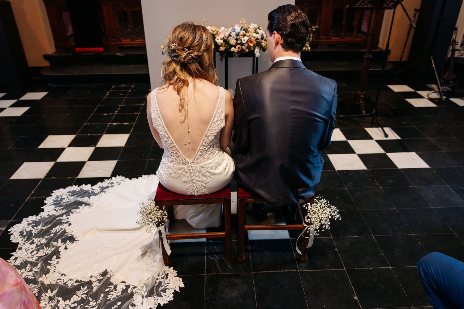 Bride and groom seated from behind at church altar during wedding ceremony on checkered floor.