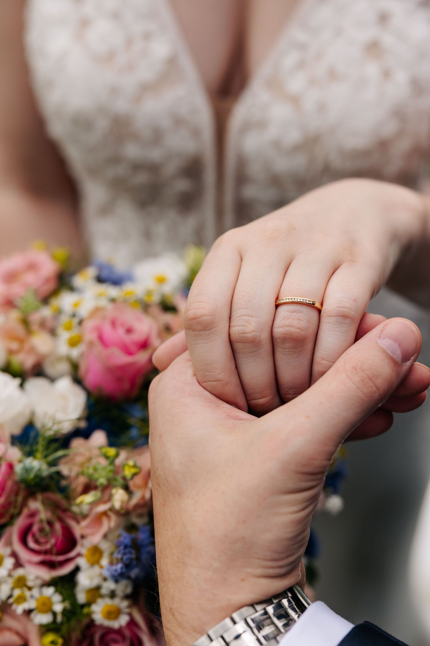 Groom holding bride's hand showing gold wedding ring with colorful floral bouquet in background.