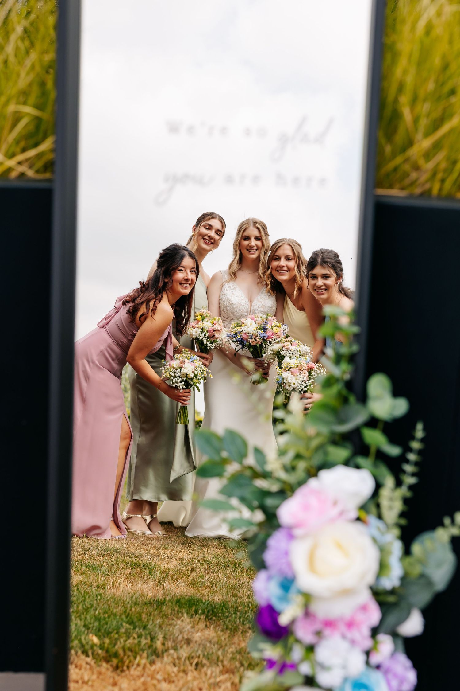 Bride and bridesmaids reflected in a decorative mirror sign surrounded by fresh flowers at an outdoor wedding.