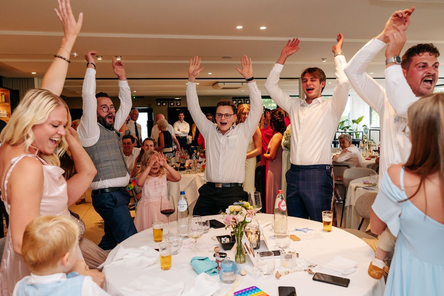Excited wedding guests raise hands and celebrate at round reception table.