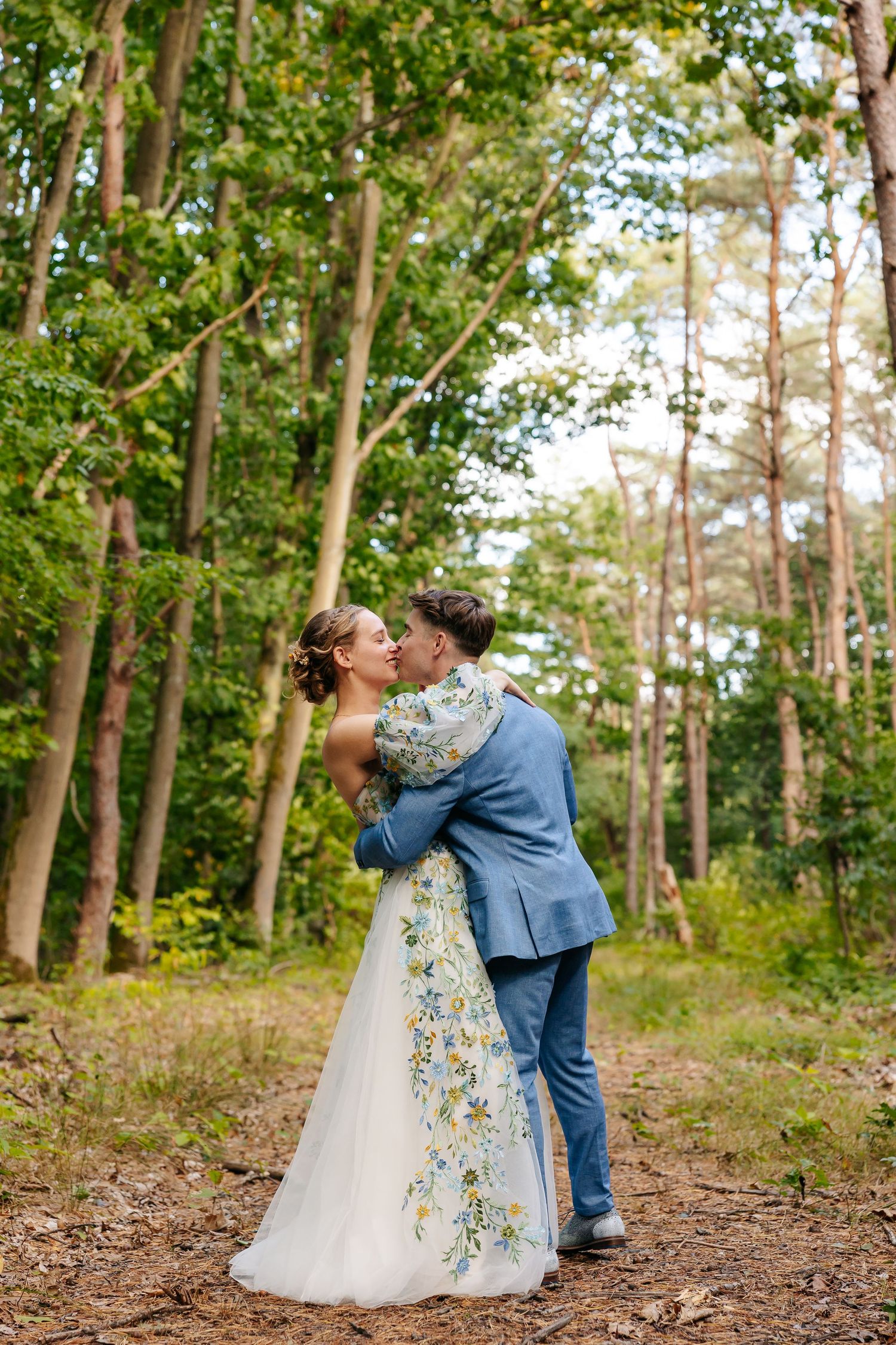 Bride and groom sharing a romantic kiss surrounded by tall trees in a lush green forest.