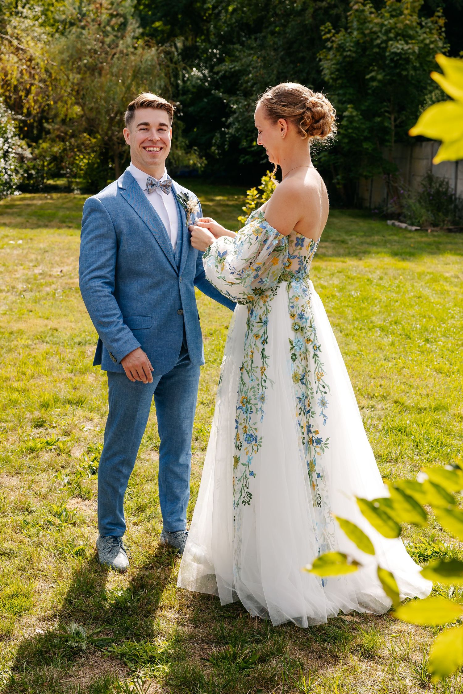 Bride in floral wedding dress adjusts grooms boutonniere in sunny garden setting.