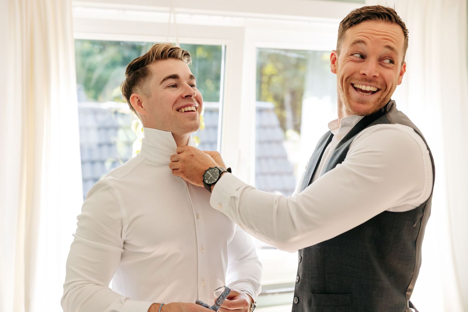 Two men laughing as groomsman adjusts groom's collar during wedding preparations.