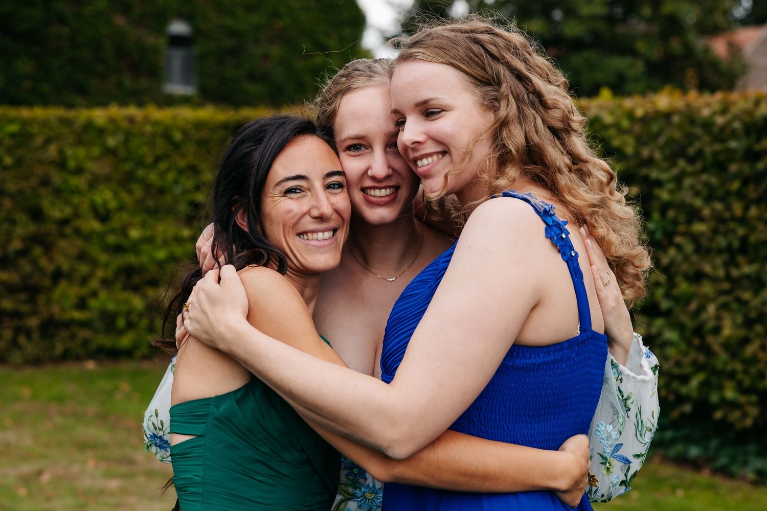 Bride embracing two smiling bridesmaids in green and blue dresses in outdoor garden setting.