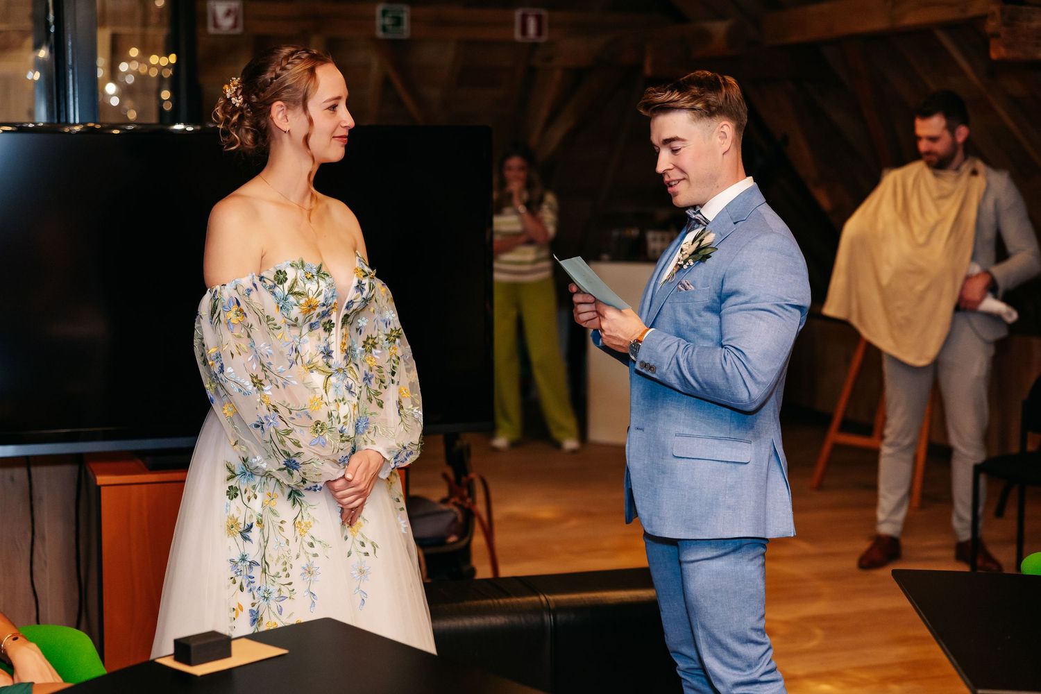 Bride in floral dress and groom in blue suit exchange vows during indoor wedding ceremony.