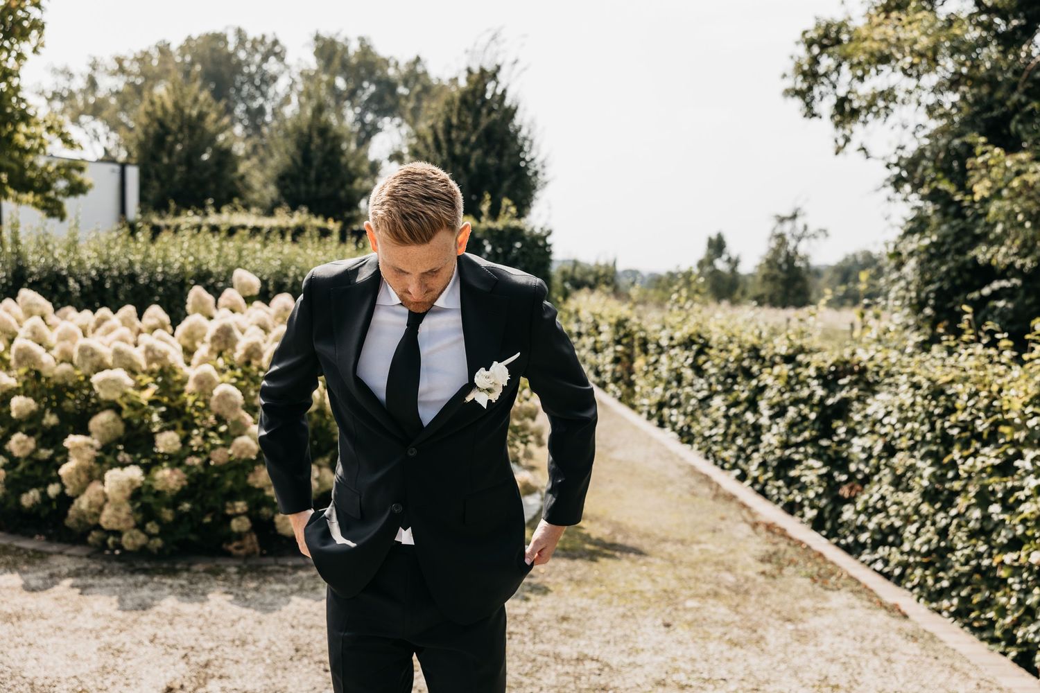 Groom in black suit with white boutonniere looking down in a garden with hydrangeas.
