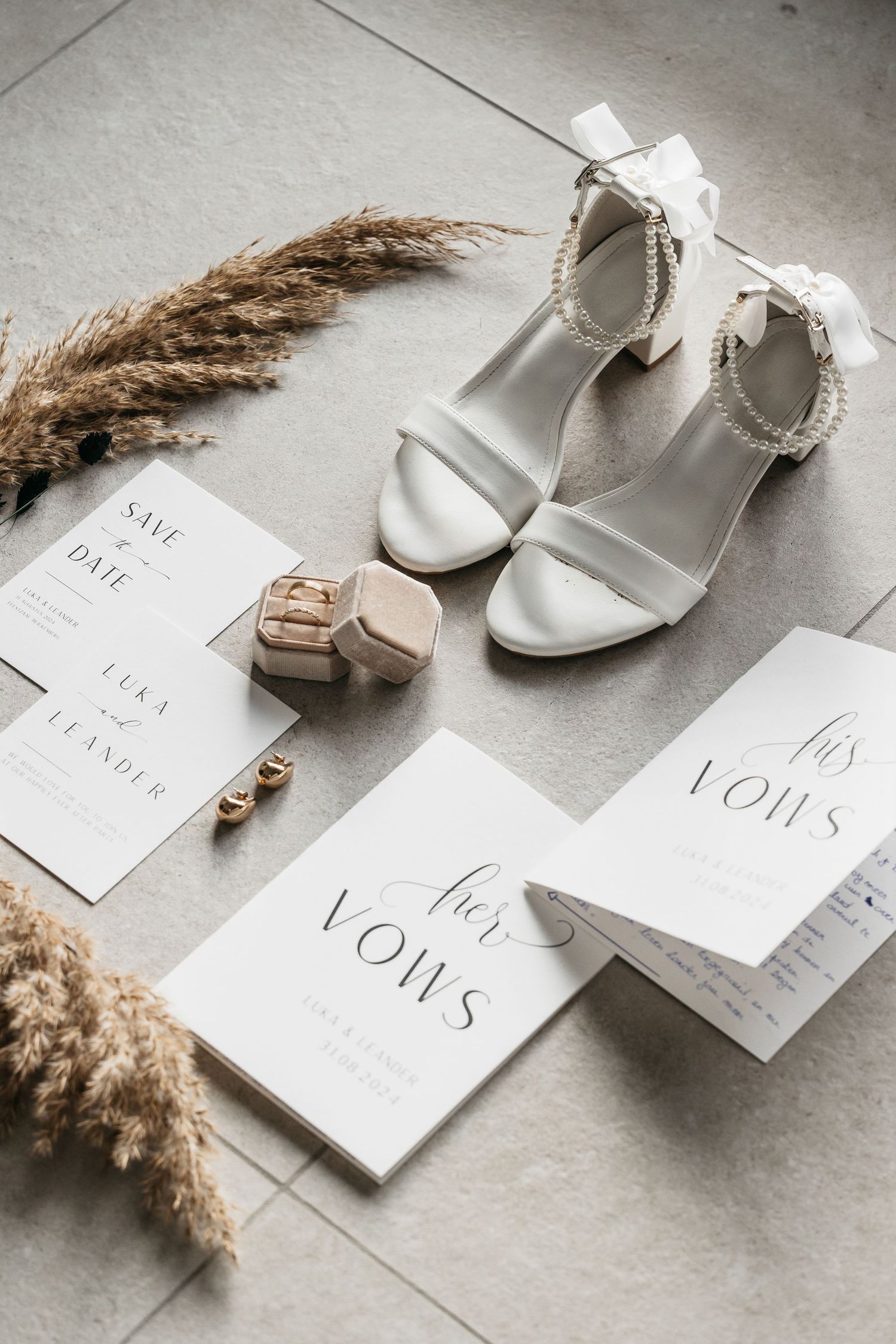 Wedding detail flat lay with white heeled shoes, vow booklets, invitation suite and pampas grass decor.