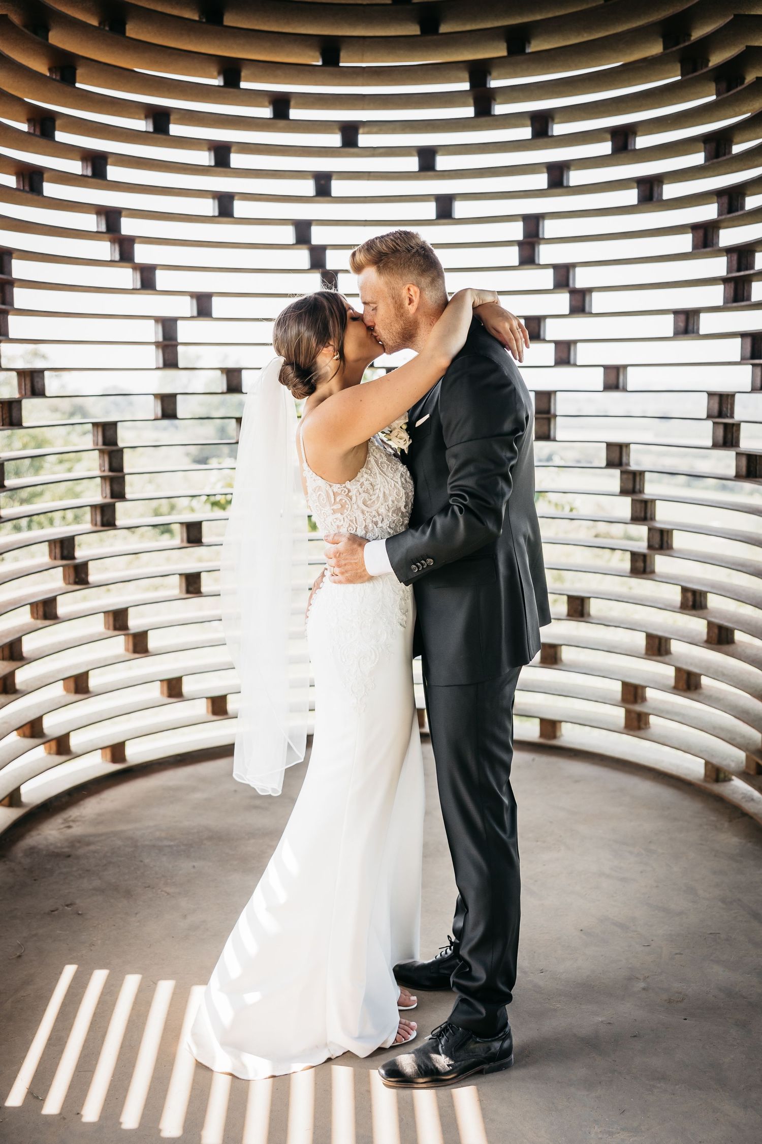 Bride and groom sharing a romantic kiss inside a curved architectural structure with patterned walls.