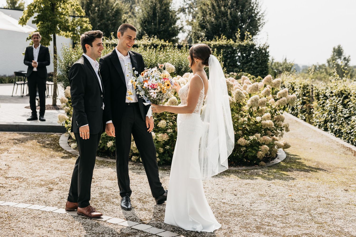 Bride in white gown with veil greets two groomsmen in black suits in a sunlit garden.