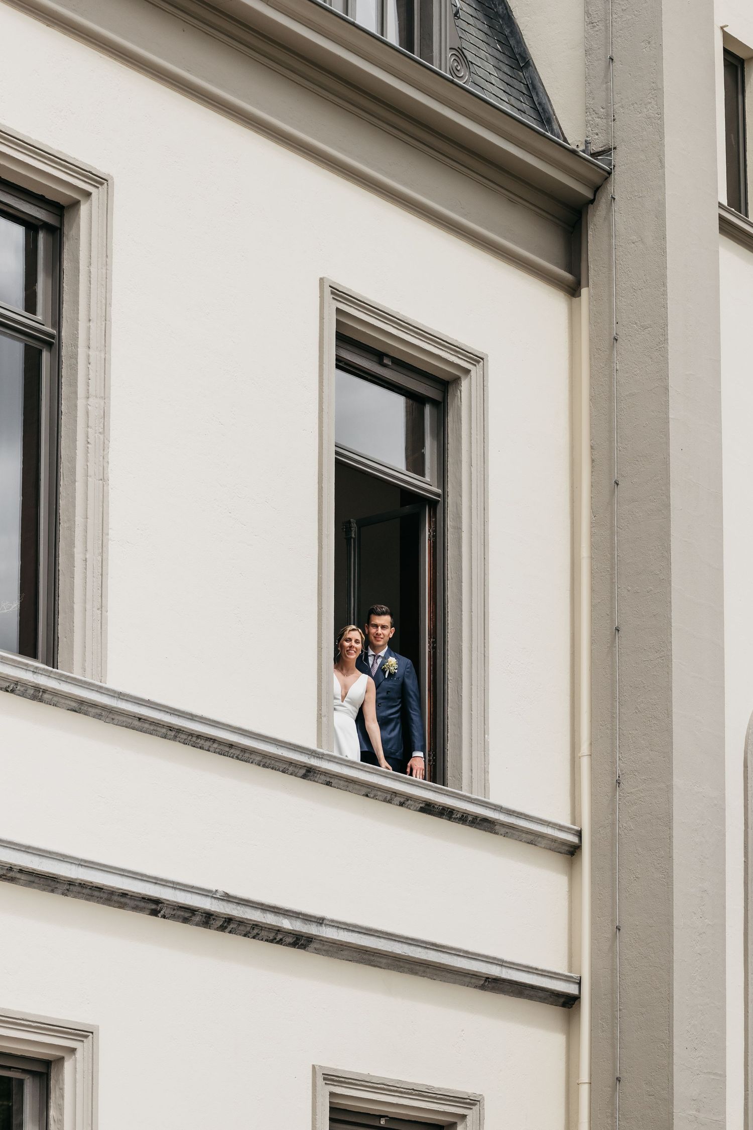 Bride in white gown and groom in navy suit pose in open window of a grand white classical building.