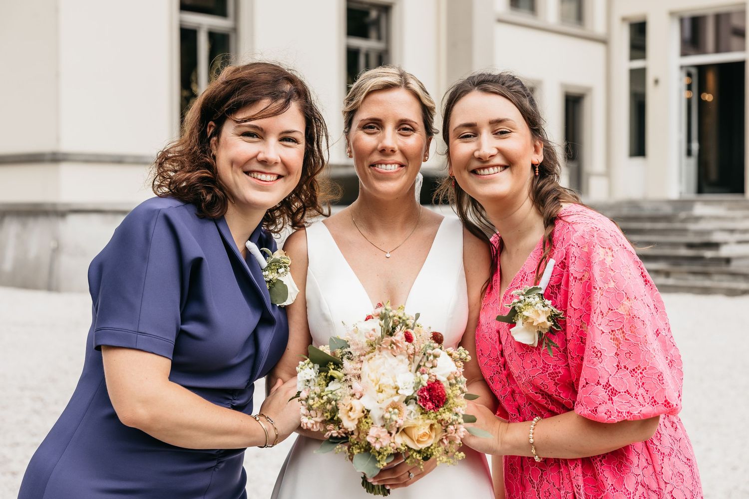 Bride in white gown smiles with two female guests in navy and pink dresses holding bouquets outdoors.