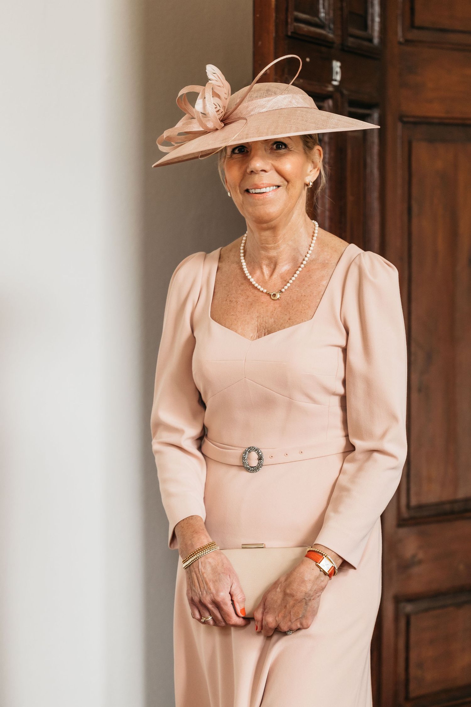 Elegant older woman in blush pink dress and matching fascinator hat at wedding.