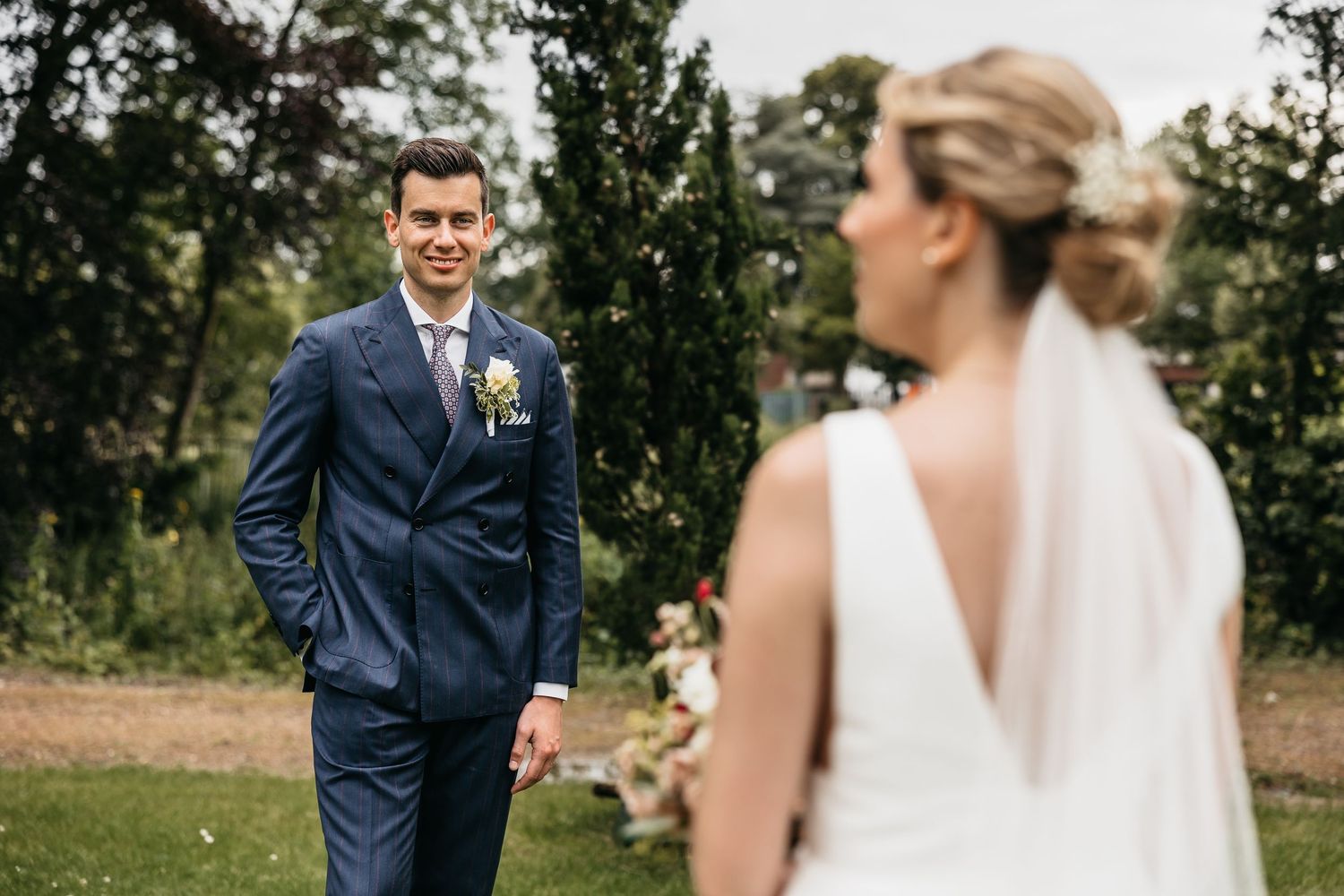 Groom in navy suit gazes lovingly at bride in white dress during outdoor wedding ceremony.