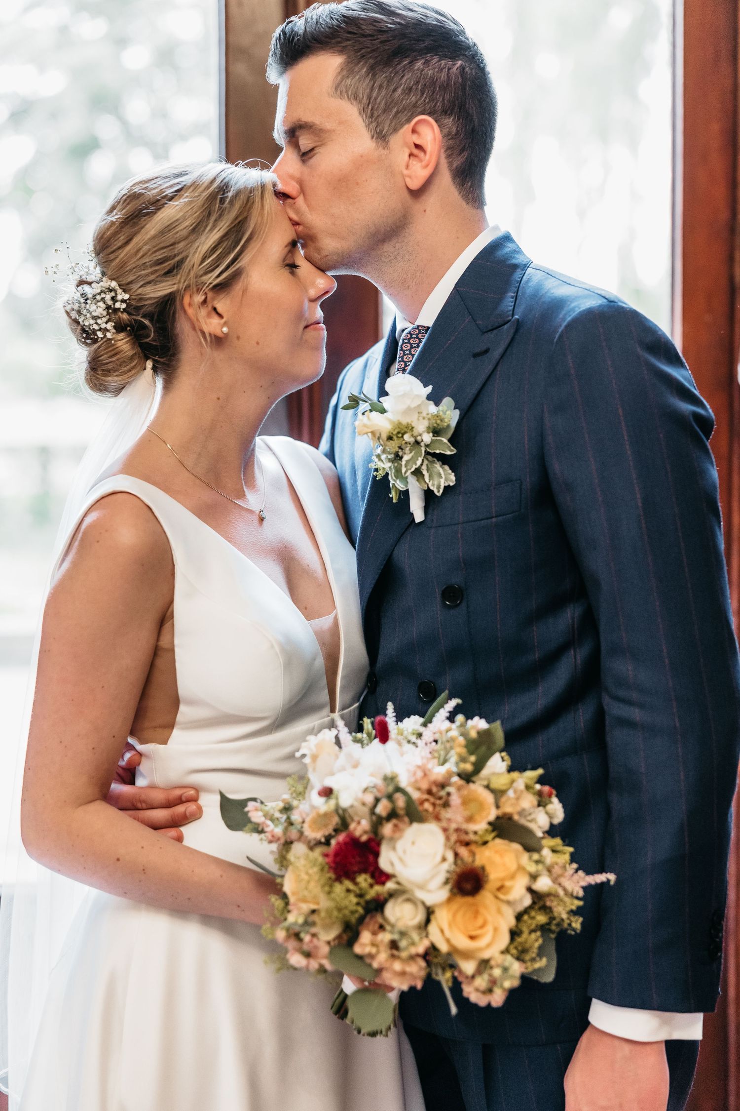 Groom kissing bride on forehead while she holds colorful wedding bouquet, standing near bright window.