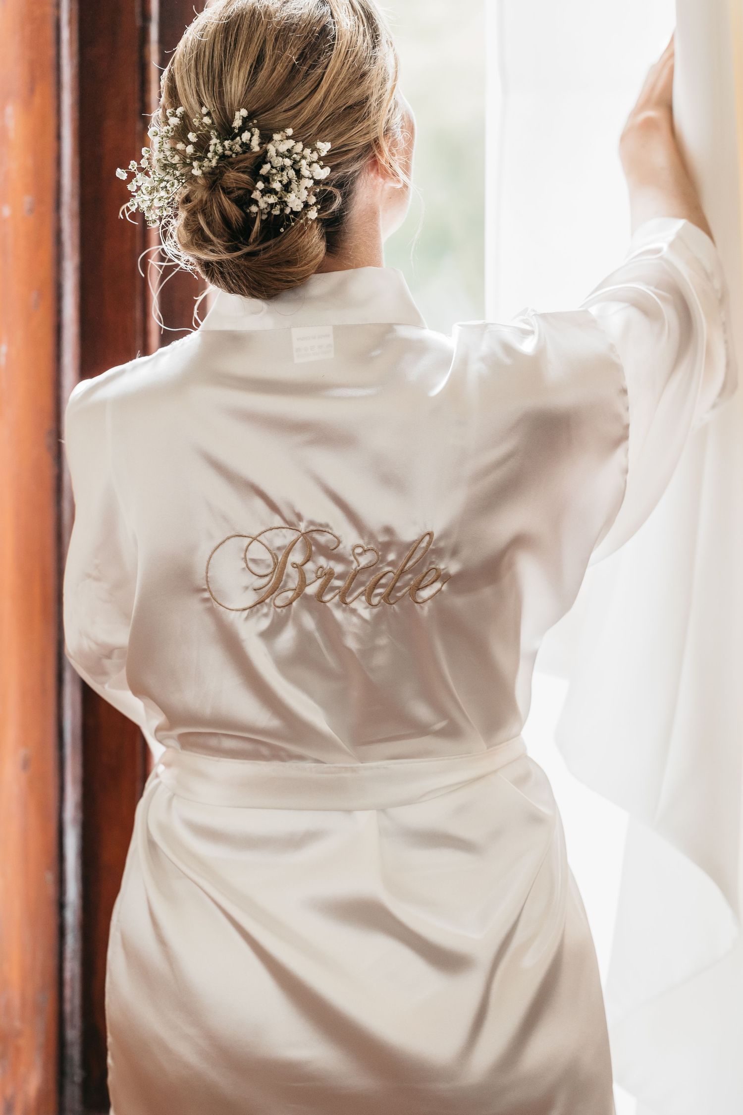 Bride wearing a white satin robe with gold embroidered text, floral hair accessory, standing by a window.