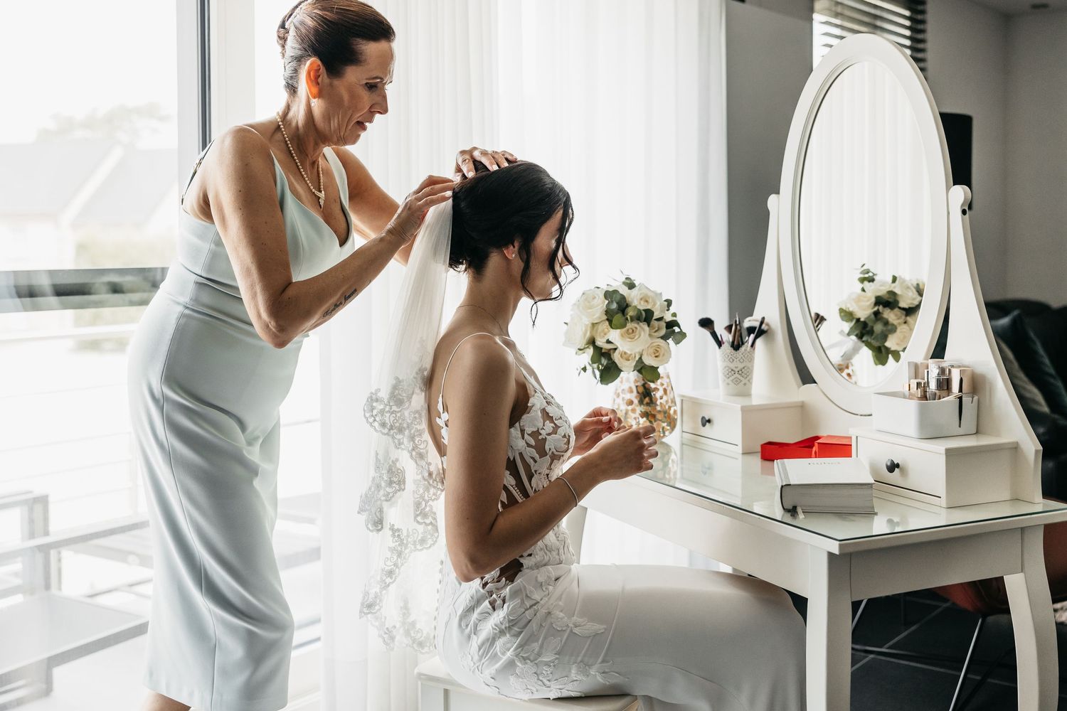Bridesmaid helping bride with veil at white vanity table during bridal preparations on wedding day.