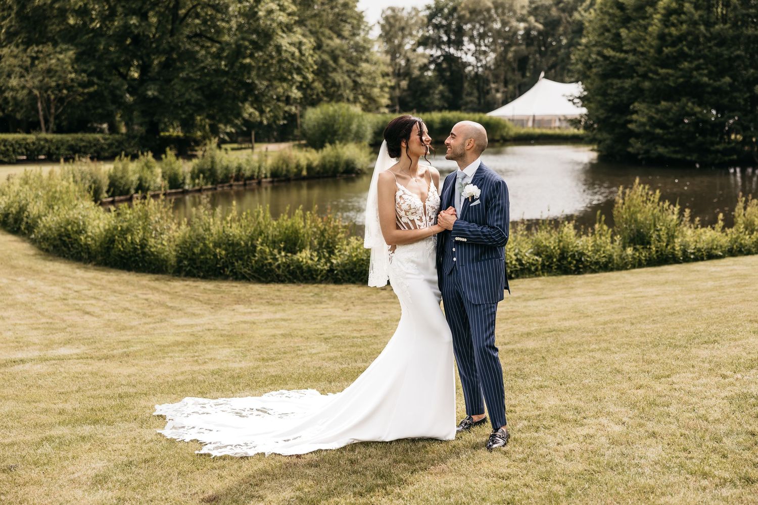 Bride and groom share a tender moment on a manicured lawn beside a tranquil pond at their wedding venue.