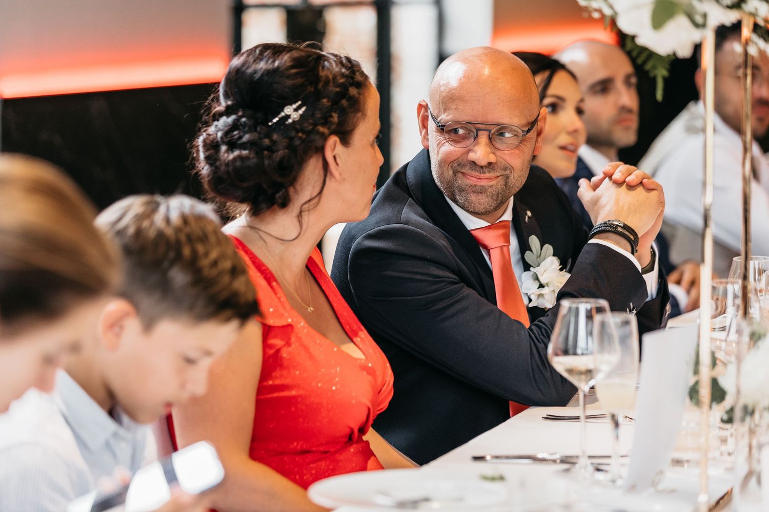 Wedding guest in navy suit and glasses smiles warmly during elegant reception dinner celebration.