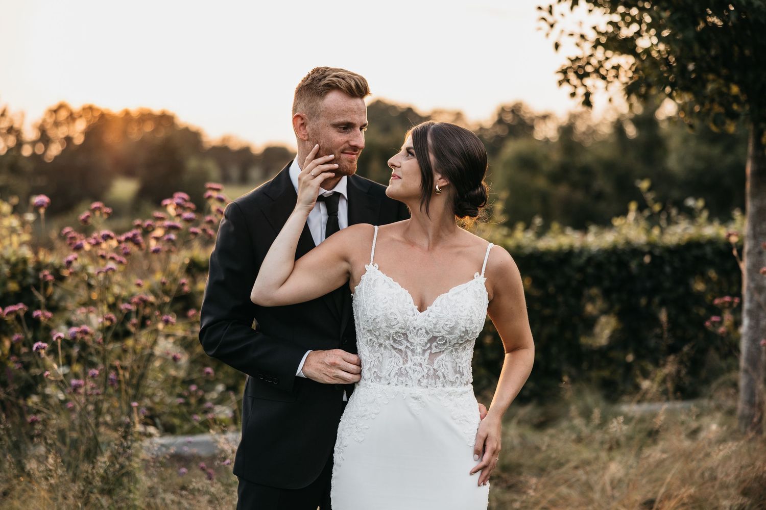 Bride in white lace spaghetti strap gown walks alongside groom in black suit through a wildflower garden at sunset.
