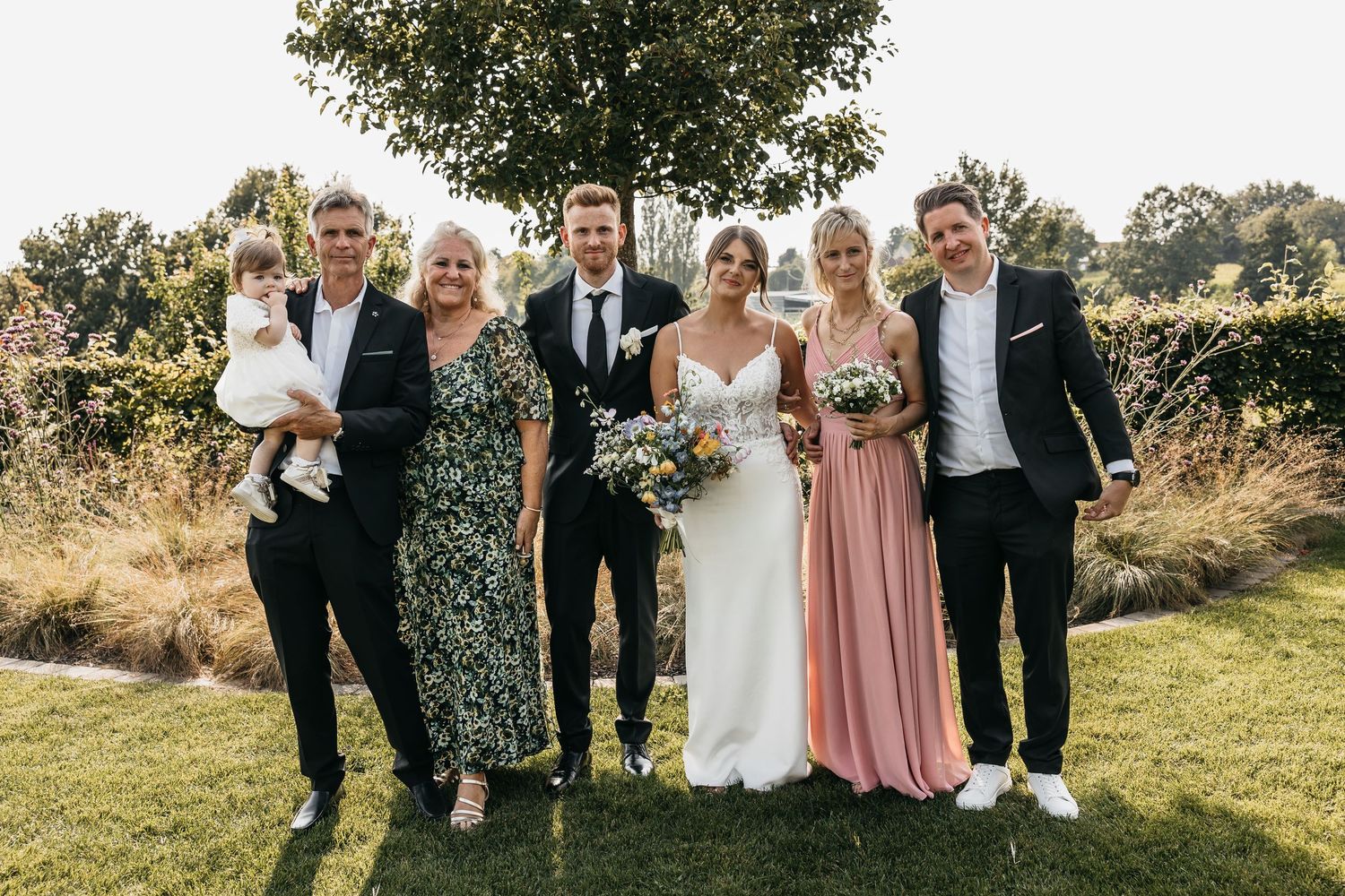 Wedding party of six posing outdoors, including bride in white gown holding colorful bouquet with groom and family members.