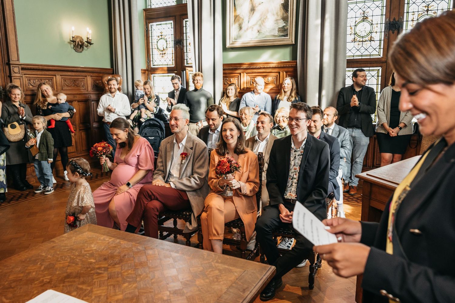 Officiant addresses smiling wedding guests seated in a beautifully decorated historic ceremony hall.