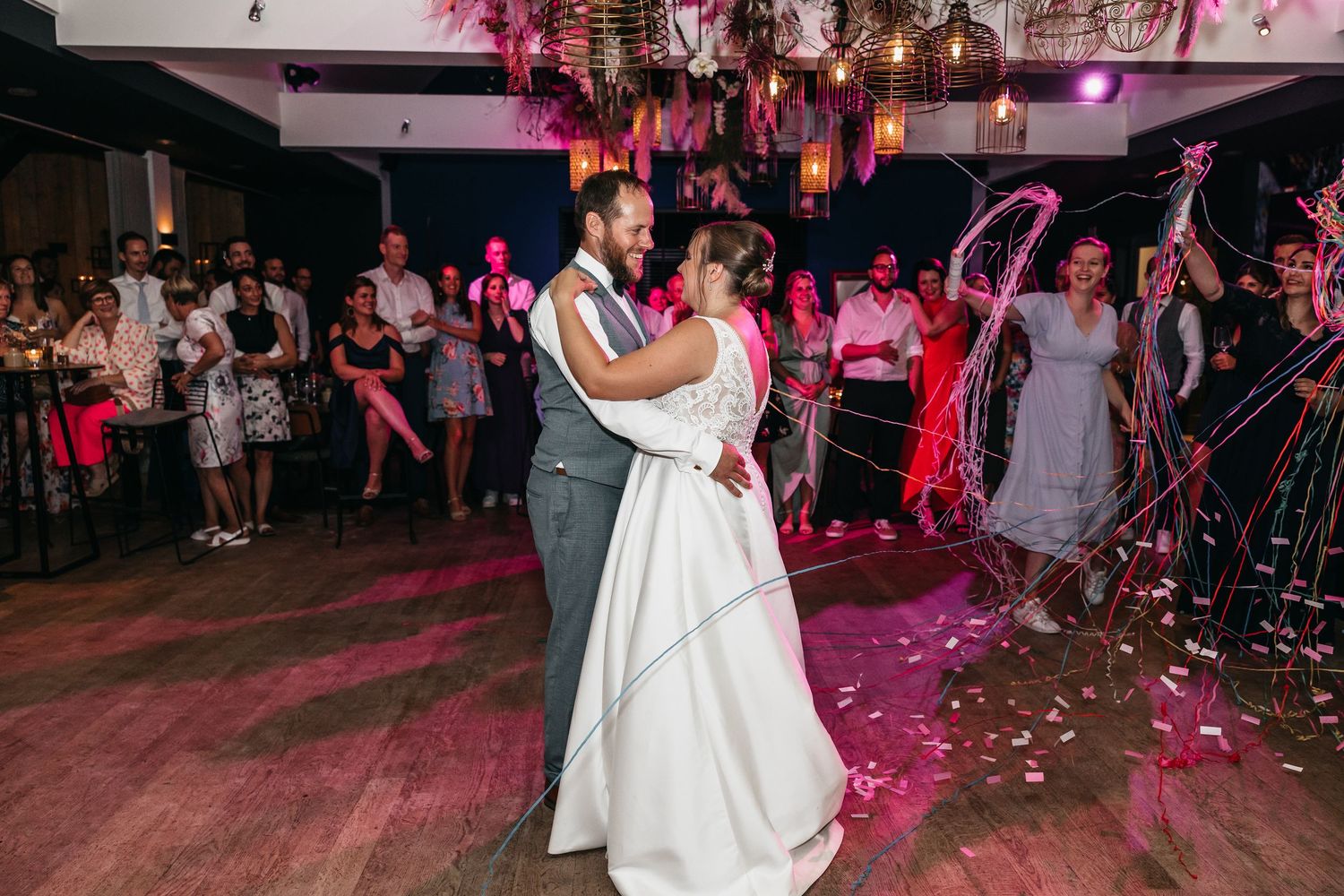 Newlyweds share a romantic first dance surrounded by guests waving streamers and pink confetti at their wedding reception.