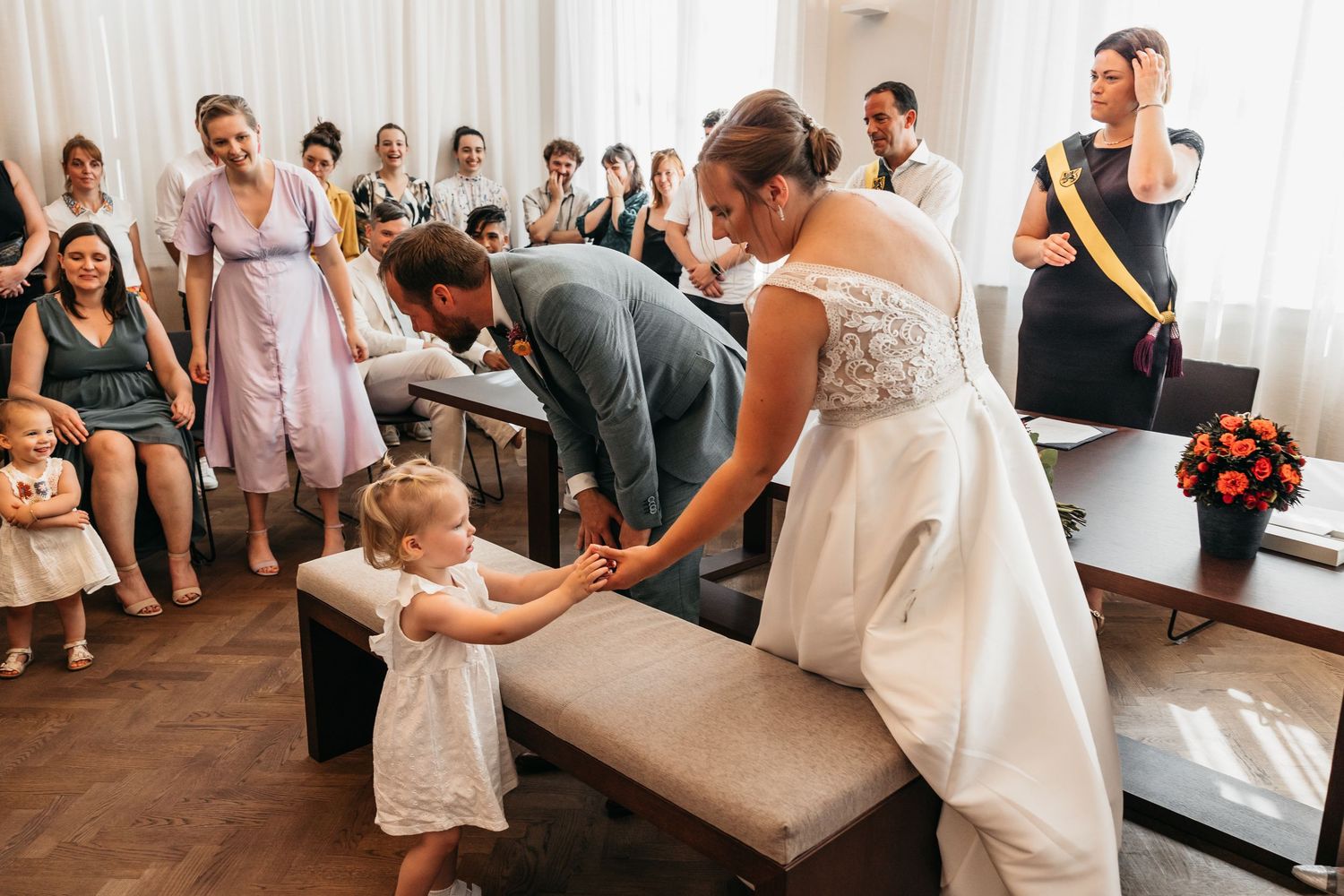 Bride and groom lean down to greet a small child in a white dress during their wedding ceremony.