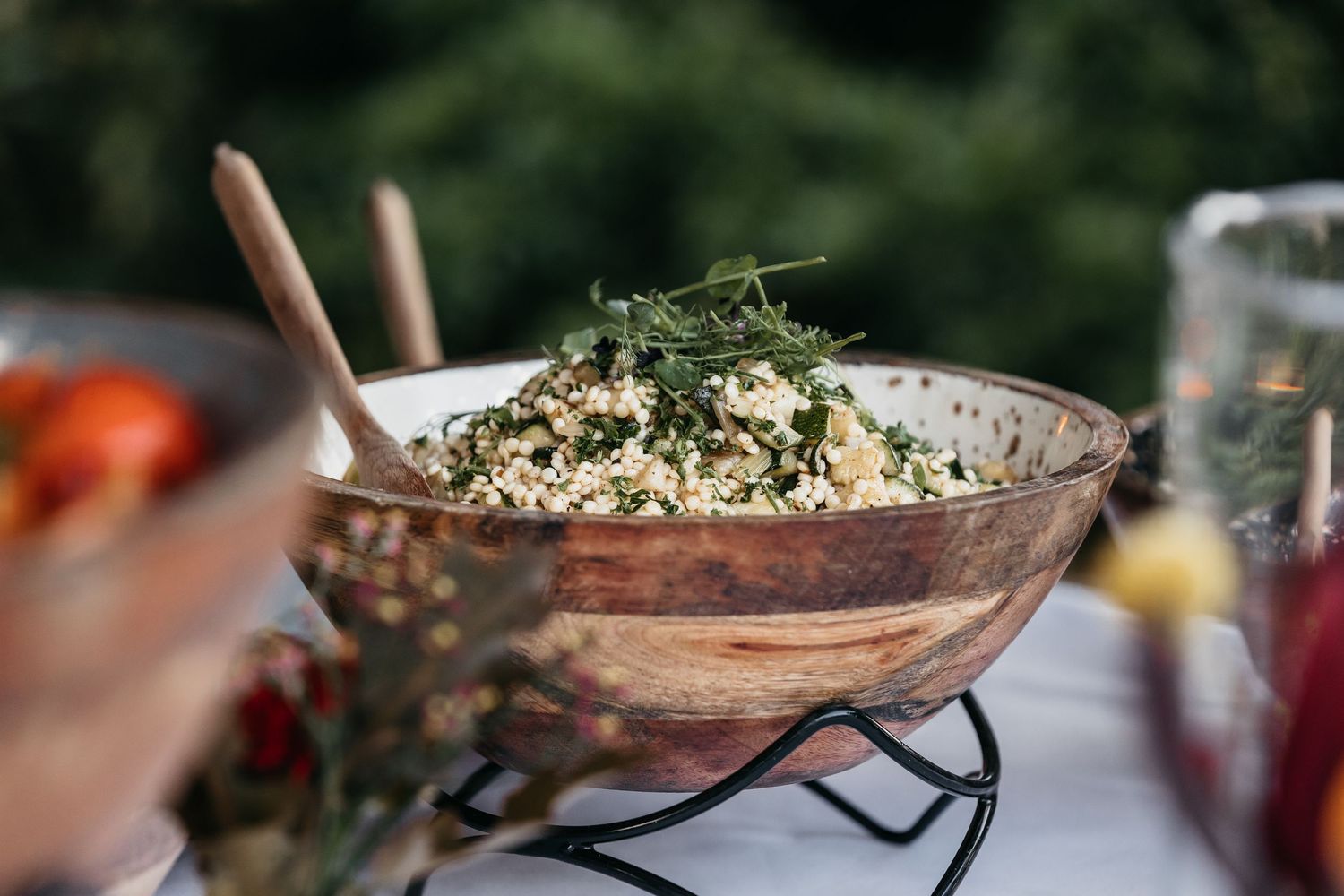 Rustic wooden bowl filled with herb and grain salad on a decorative metal stand outdoors.