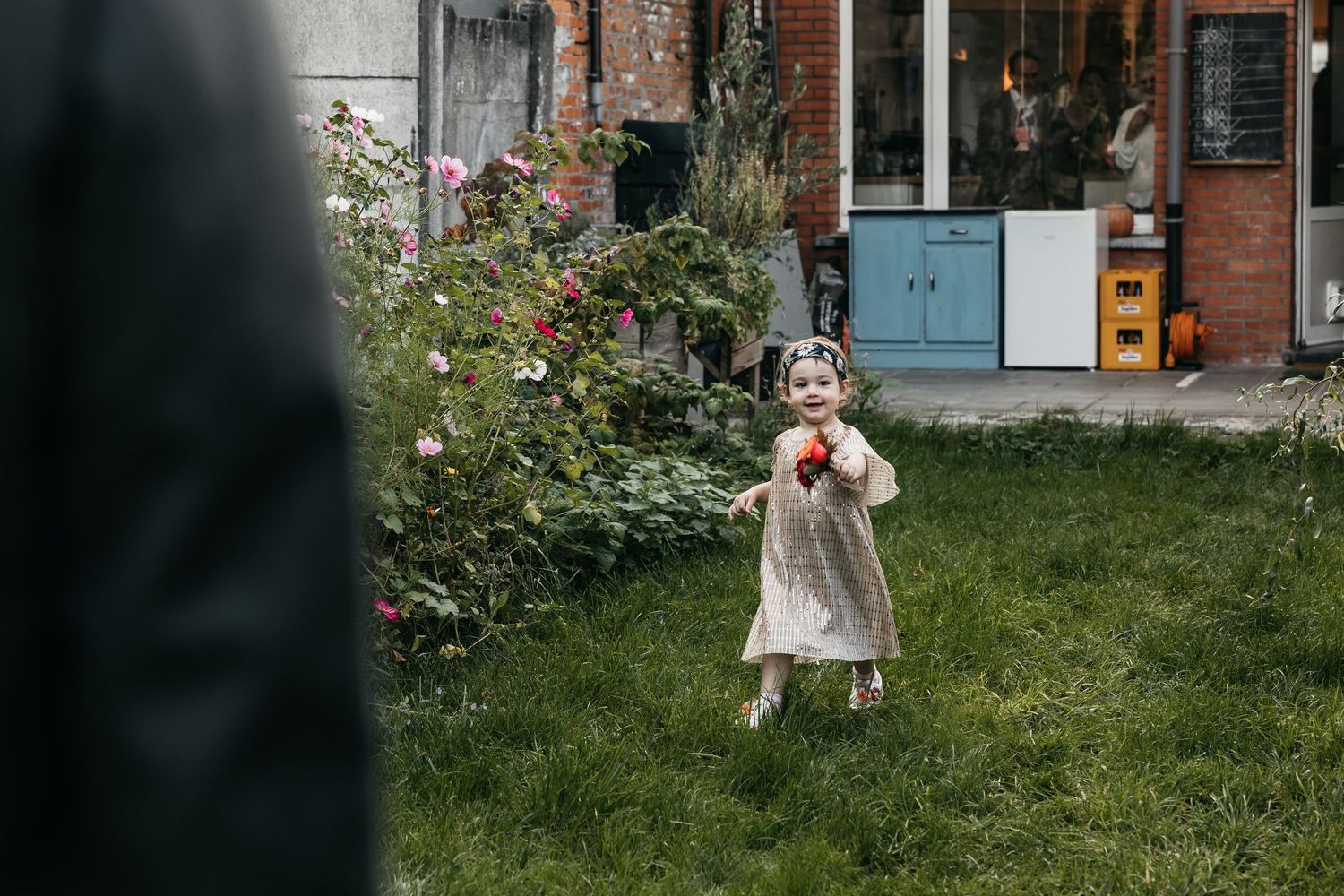 Adorable flower girl in cream dress holding a red flower in a garden during a wedding.