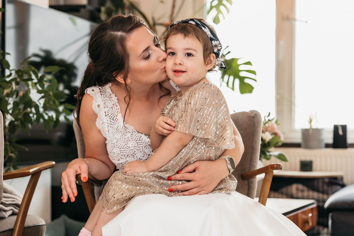 Bride in white lace dress kissing a young flower girl in a gold sequin dress on the cheek.