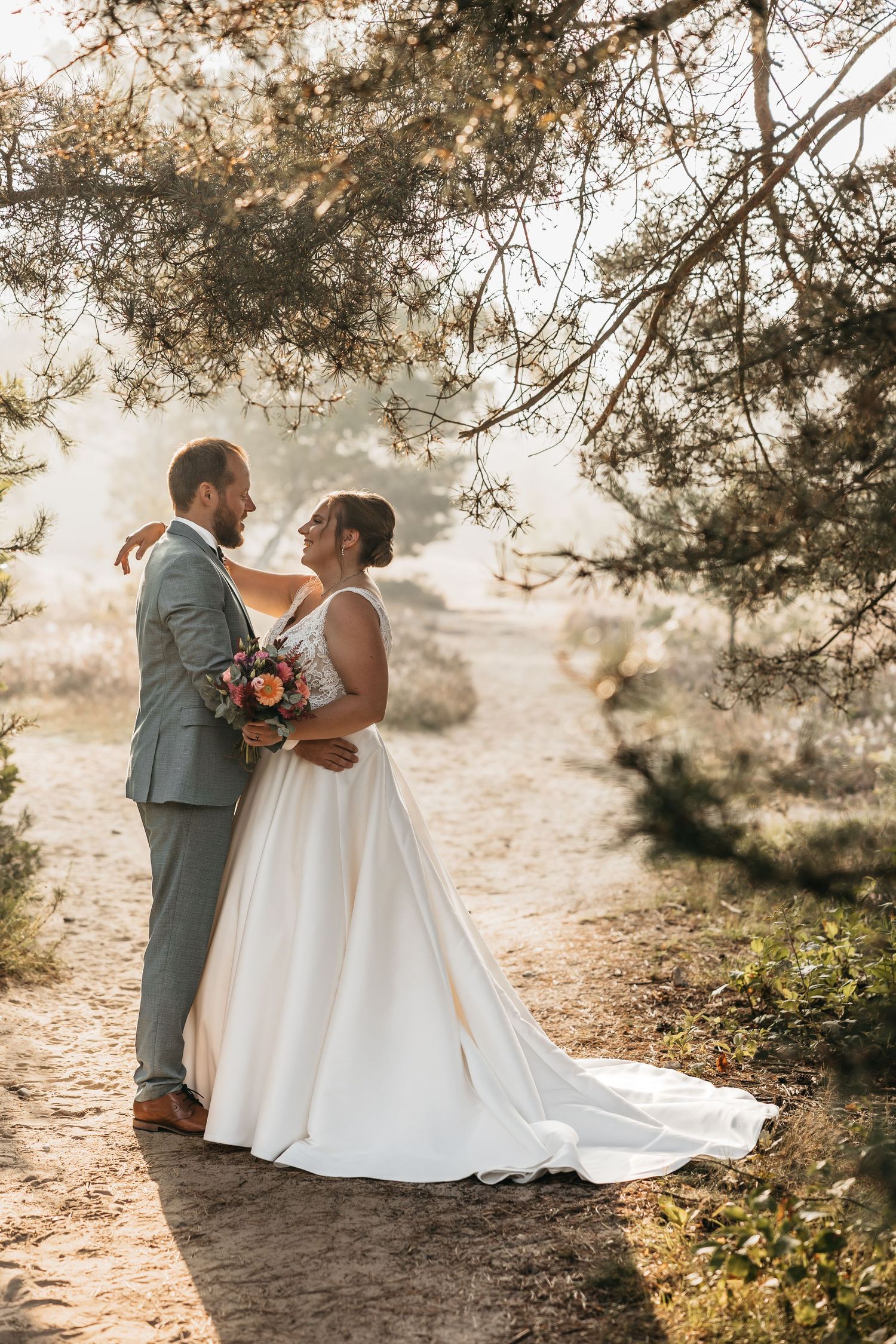 Bride in flowing white gown and groom embrace under pine tree branches in golden afternoon light.