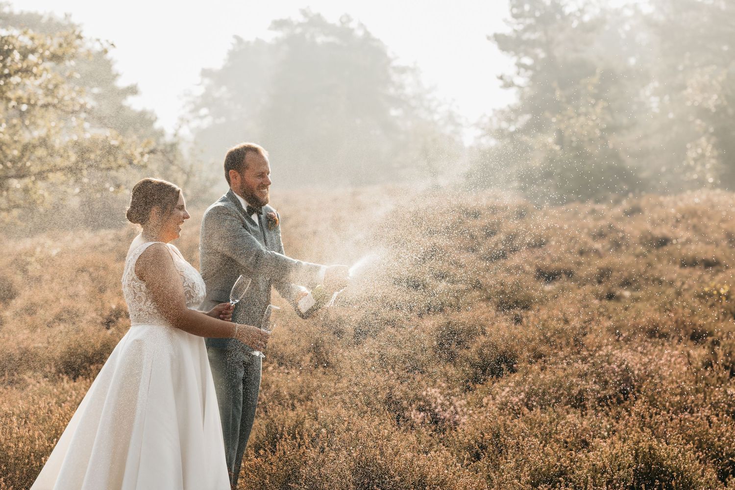 Groom popping champagne bottle as bride smiles in a warm golden outdoor heather landscape.