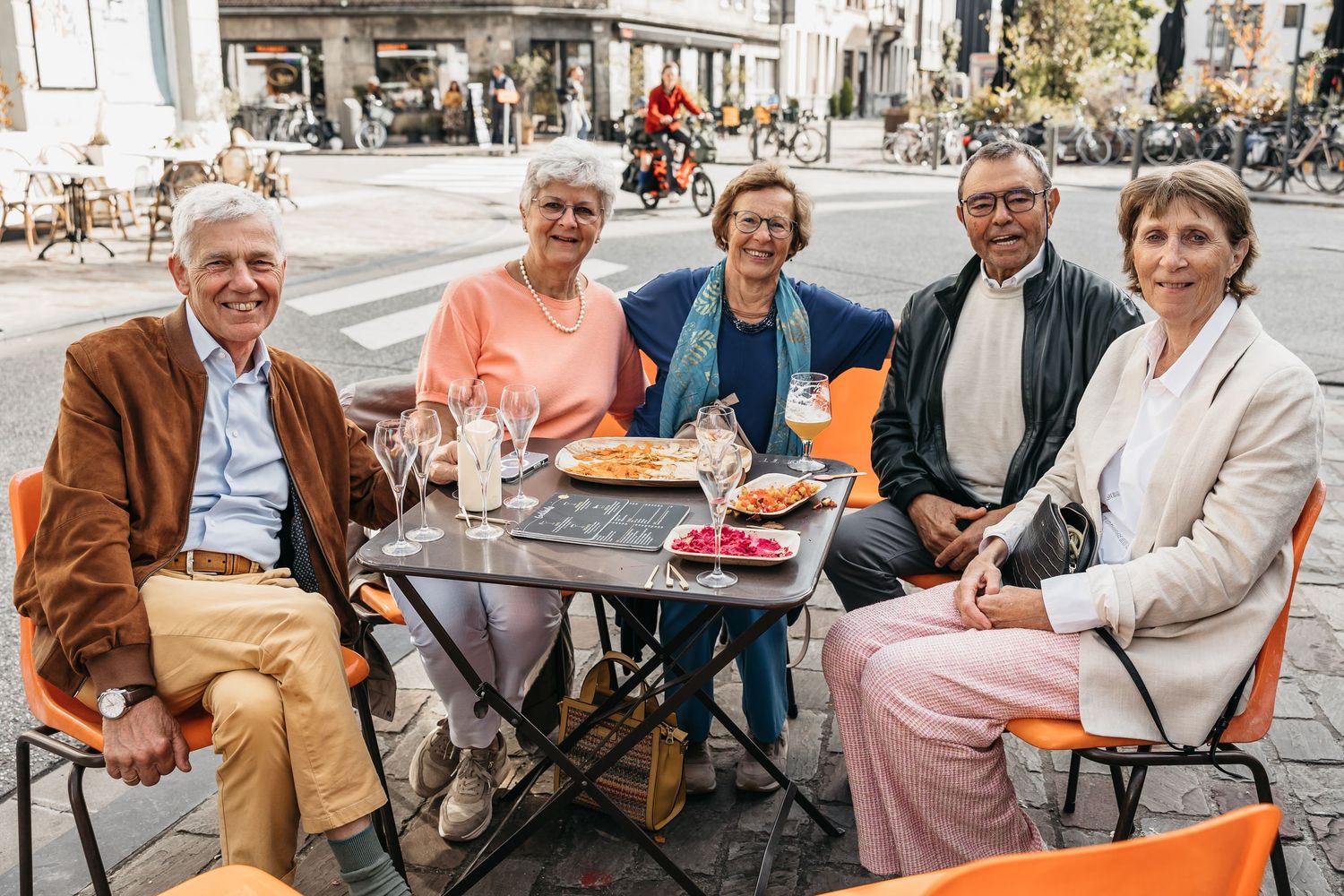 Five smiling adults sitting together at an outdoor café table on a sunny city street.