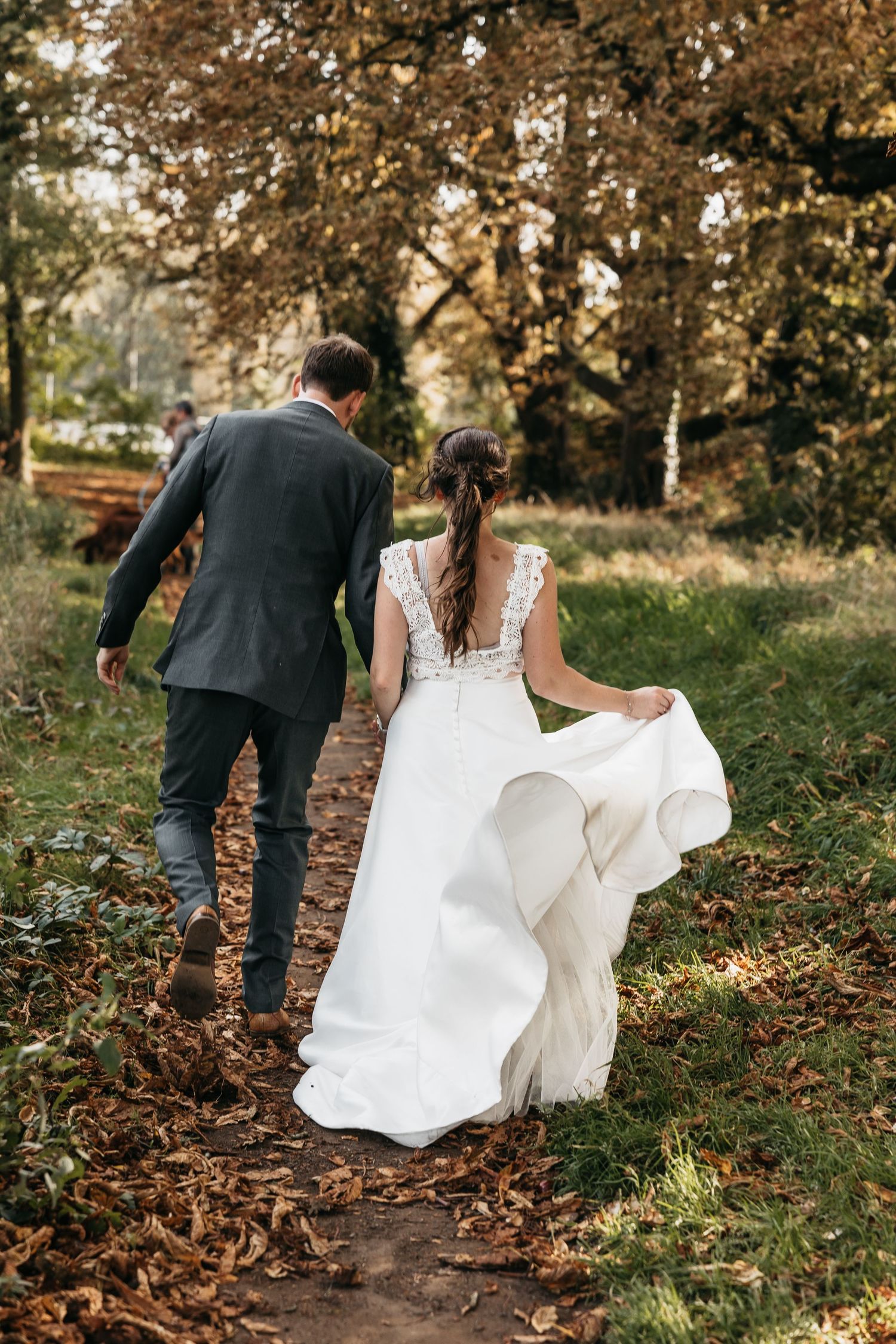 Wedding couple walking away on a leaf-covered path through golden autumn trees holding hands.