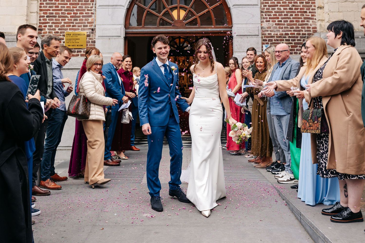 Newlywed couple walks hand-in-hand through a confetti shower outside a brick church with smiling guests.