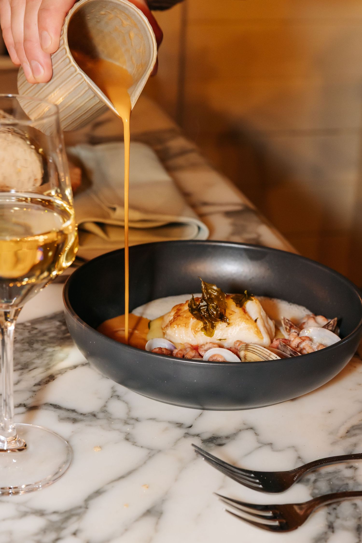 Chef pouring golden broth over seafood dish in black bowl on marble surface with white wine.