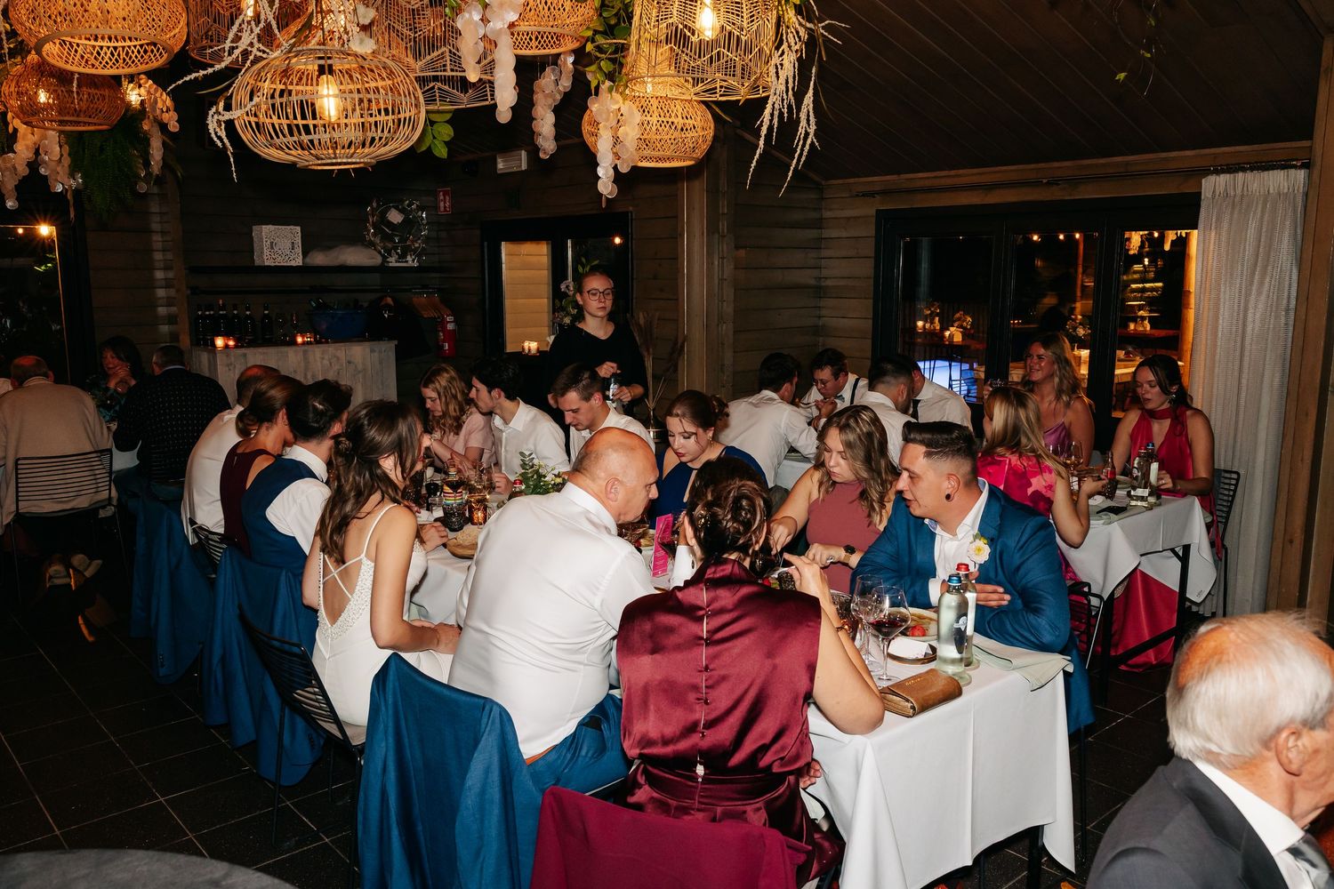 Wedding reception guests dining together at elegantly decorated tables in warmly lit rustic venue.