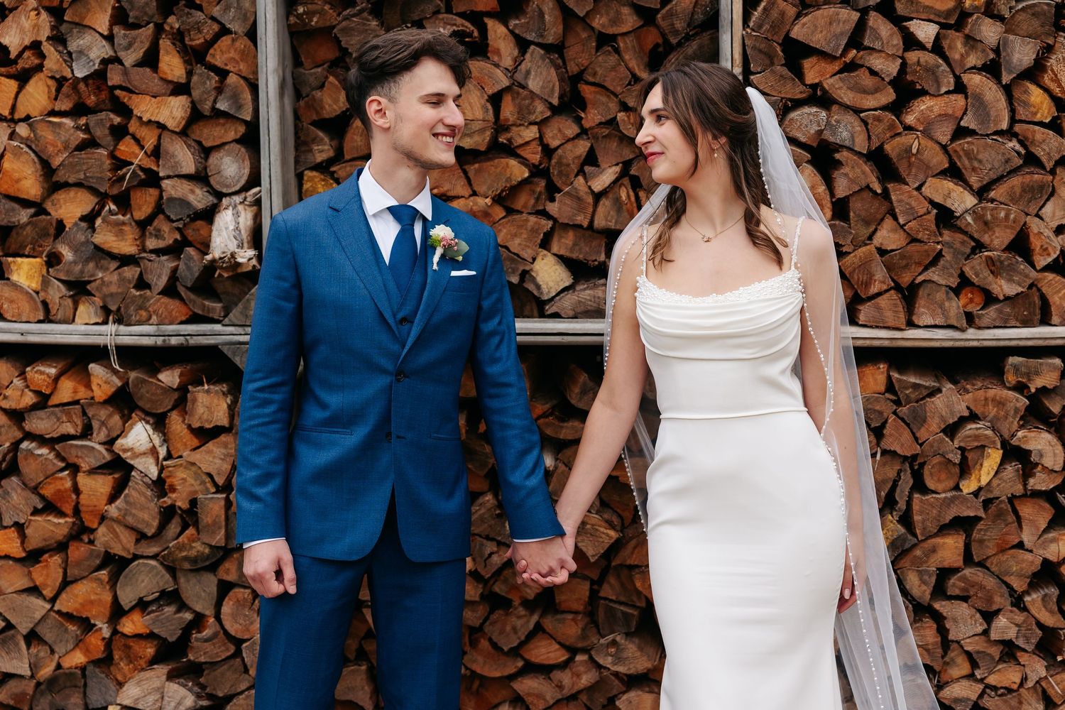 Smiling bride and groom in blue suit holding hands before a rustic stacked wood wall backdrop.