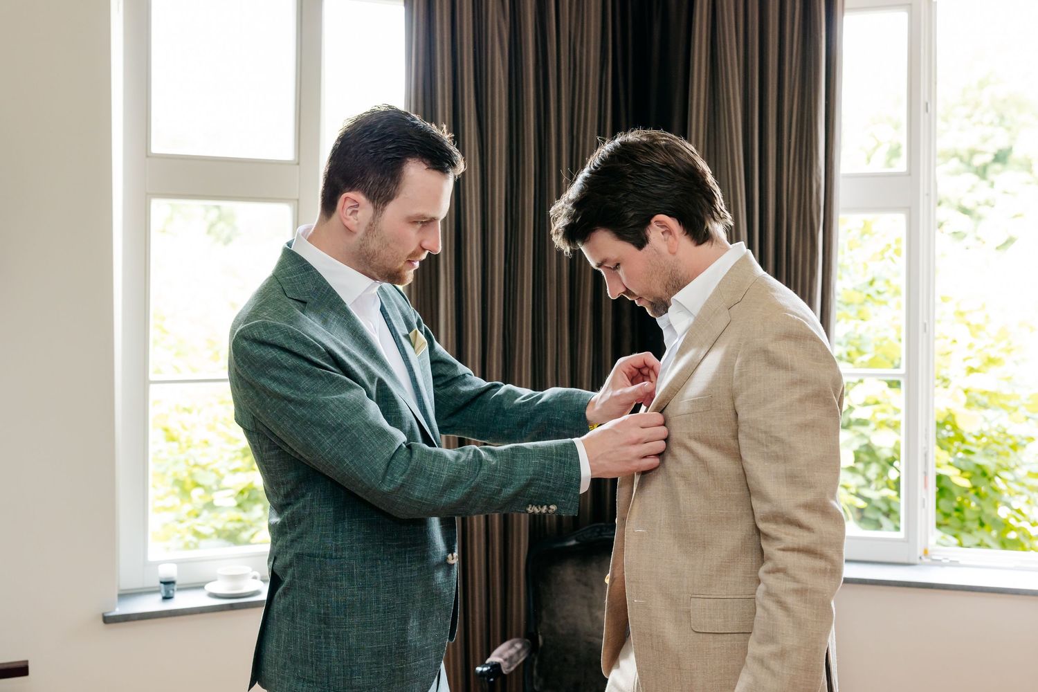 Best man helping groom adjust boutonniere on beige suit jacket before wedding ceremony.