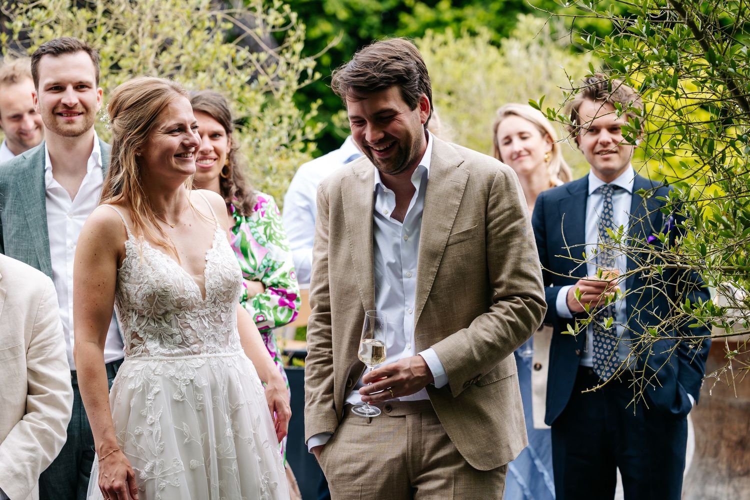 Smiling bride in lace gown and groom in tan suit mingling with wedding guests in garden setting.