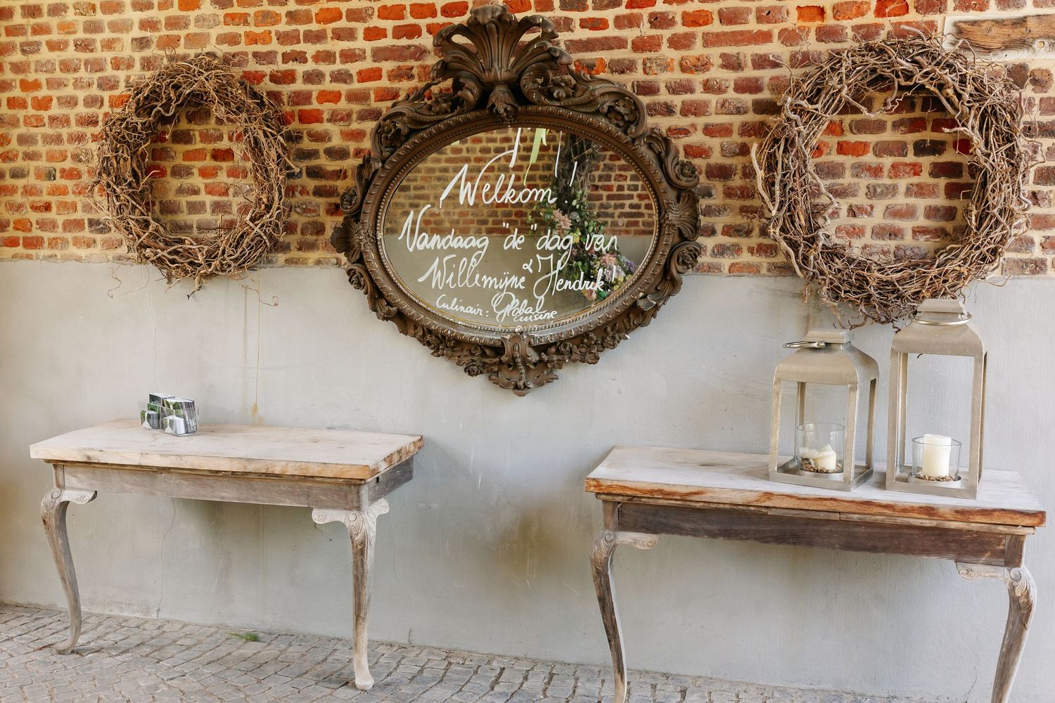 Rustic wedding welcome display with ornate mirror, twig wreaths, and elegant console tables against a brick wall.