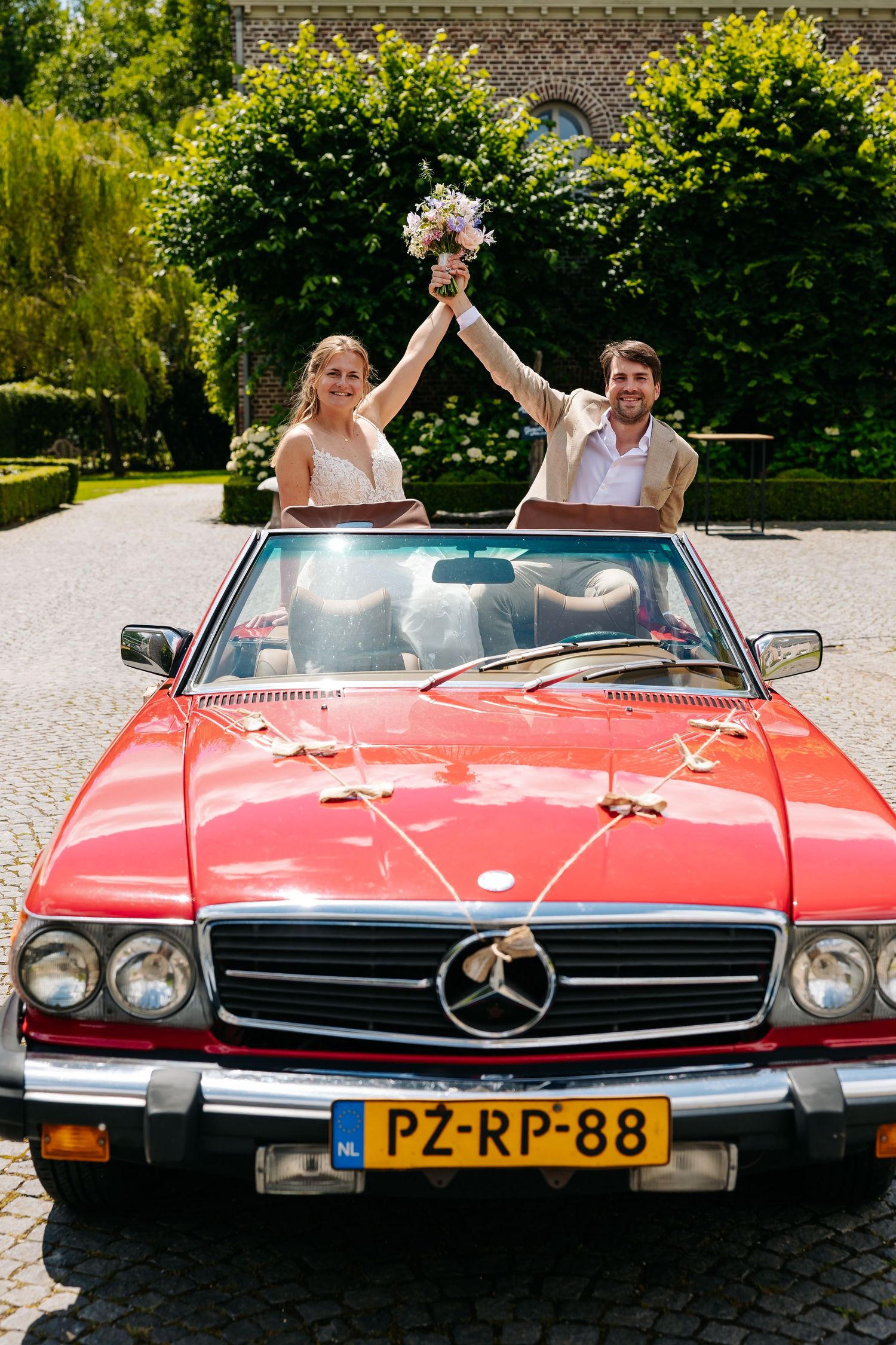 Bride and groom celebrate in classic red Mercedes convertible decorated with white ribbons at wedding.
