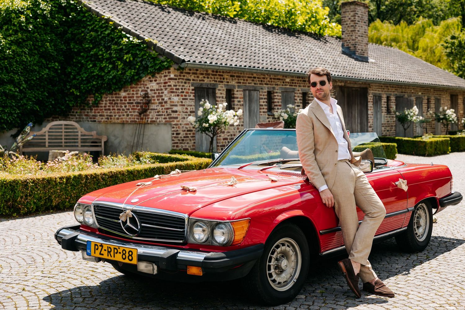 Groom in beige suit leaning against classic red Mercedes convertible outside rustic brick wedding venue.