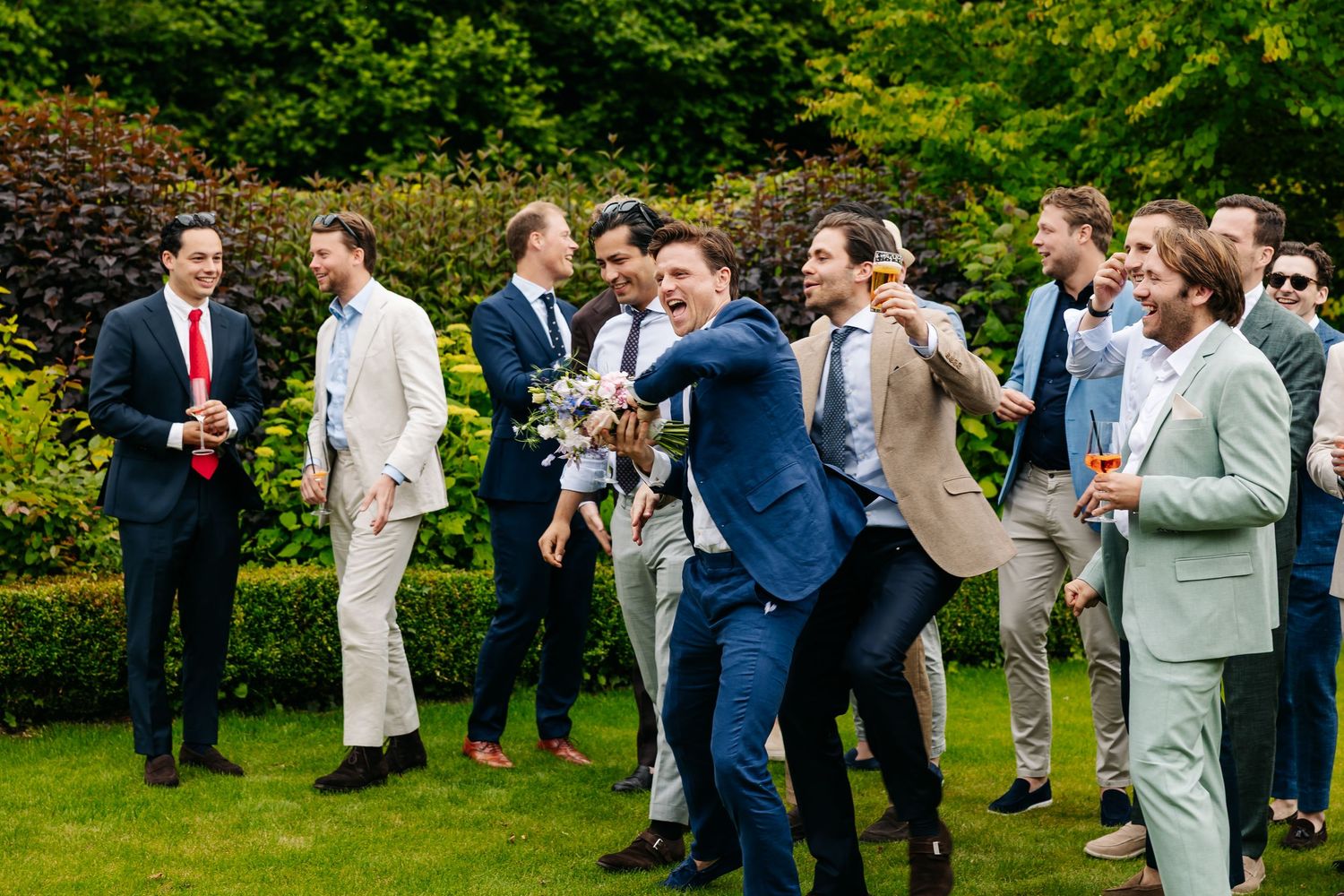 Group of men in suits laugh and dance together during outdoor wedding reception in garden.