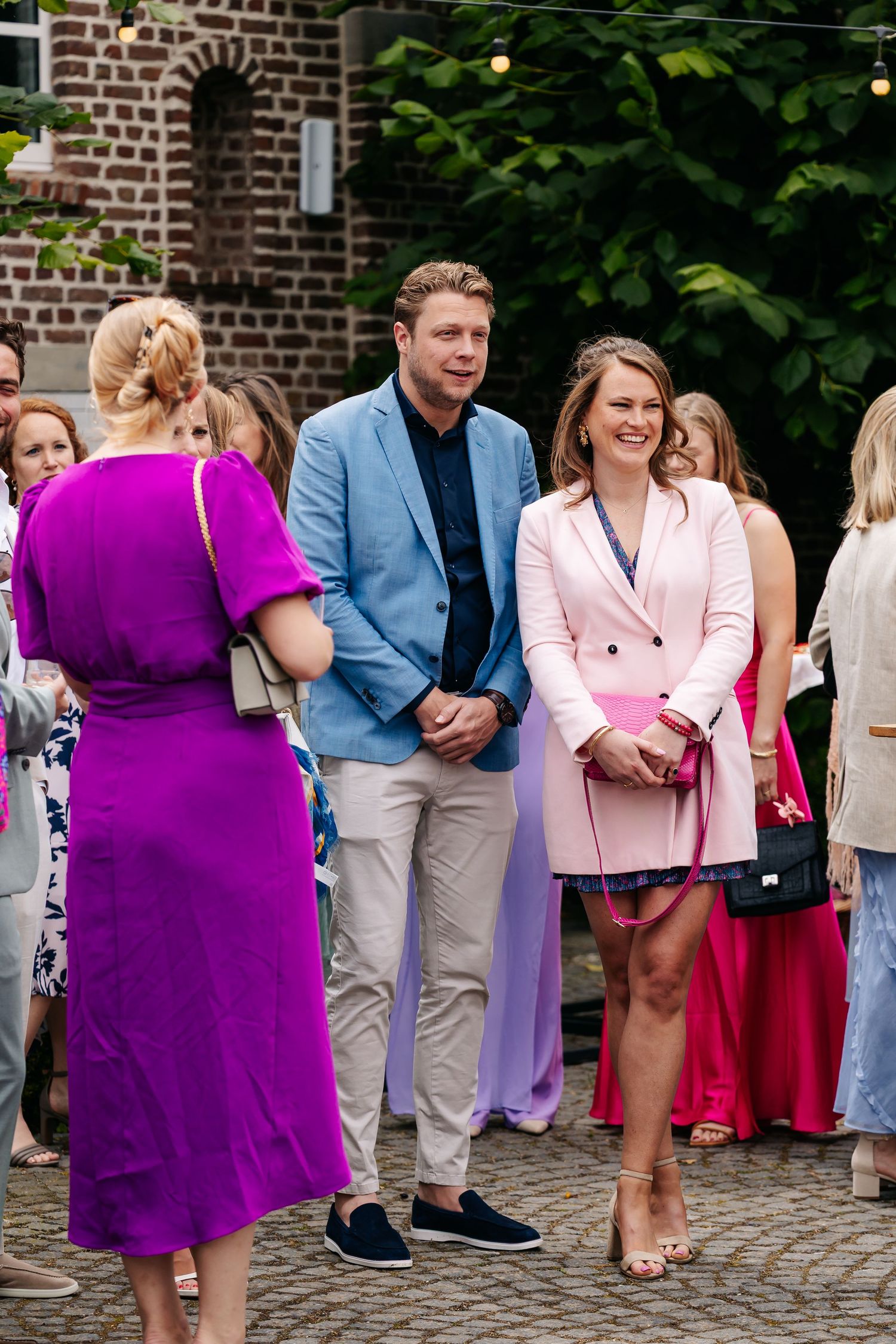 Wedding guests in colorful attire socializing outdoors, woman in pink blazer smiling with man in blue jacket.